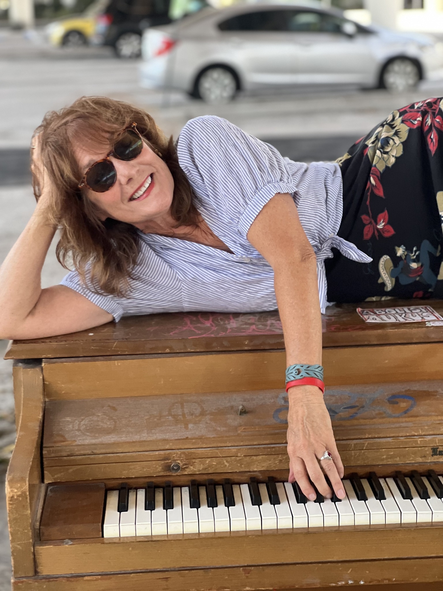 Miss Paula Bradley smiling in sunglasses, leaning across the keys of an upright piano