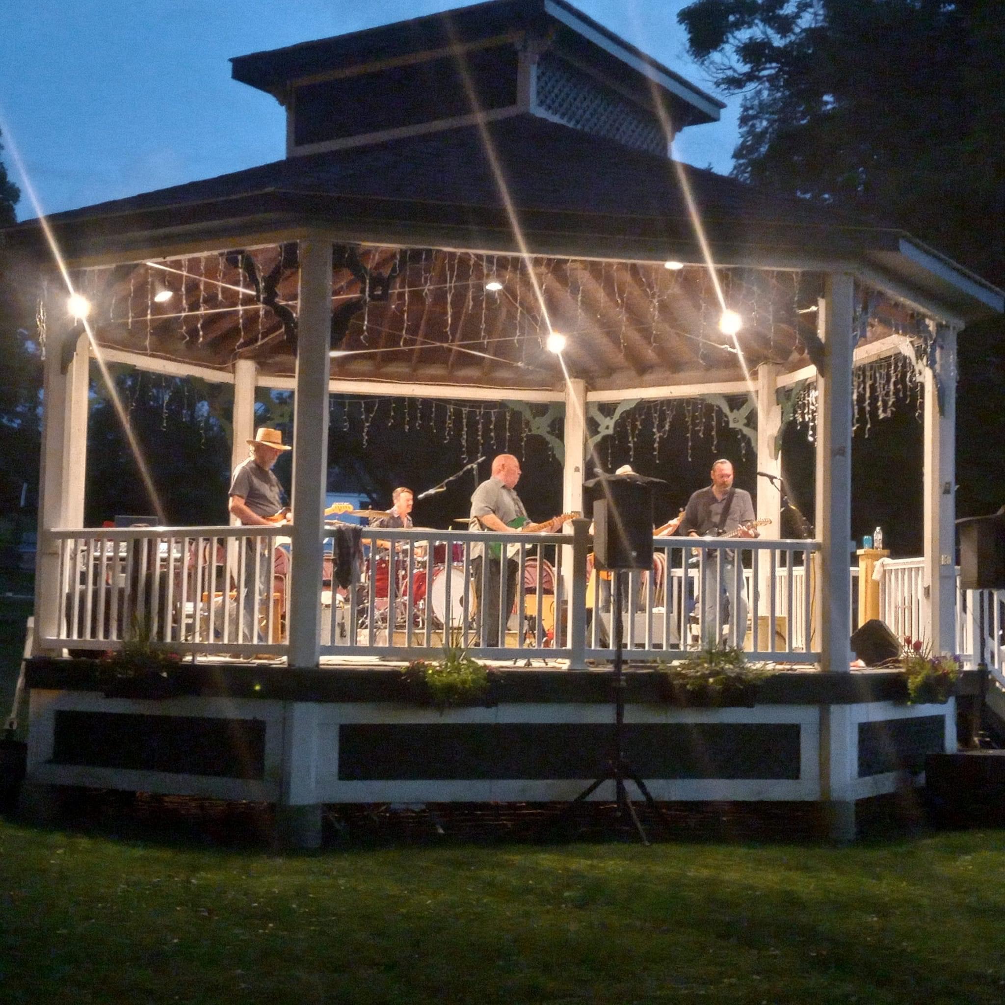 A band on the gazebo stage strung with lights as the sky turns to evening.