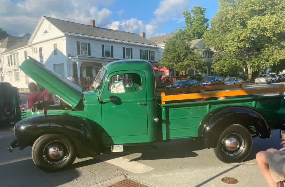 A restored green pickup with its hood raised on a village street at the car show.