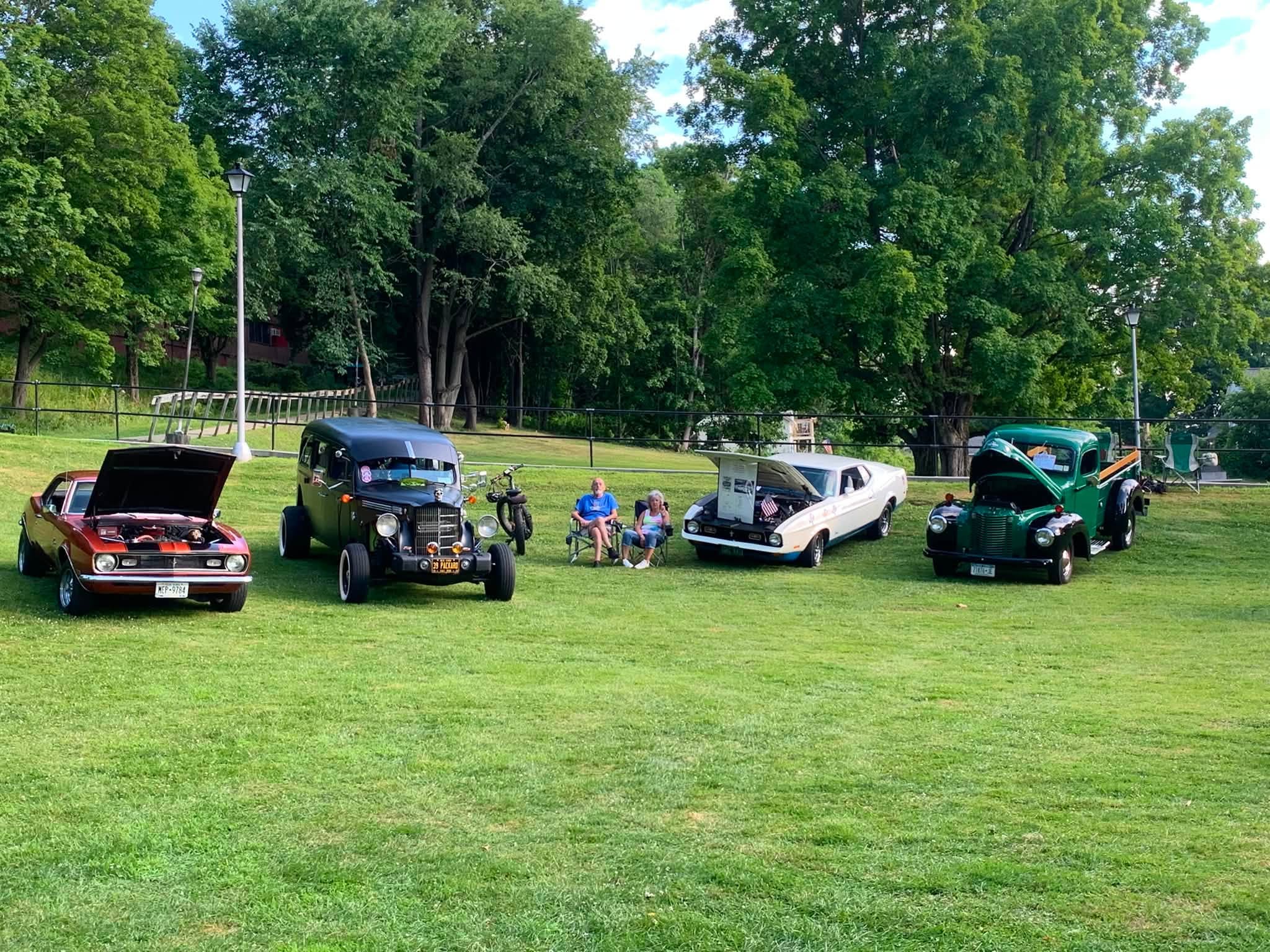 Classic cars with their hoods up across the Wood Park lawn while owners relax in lawn chairs.