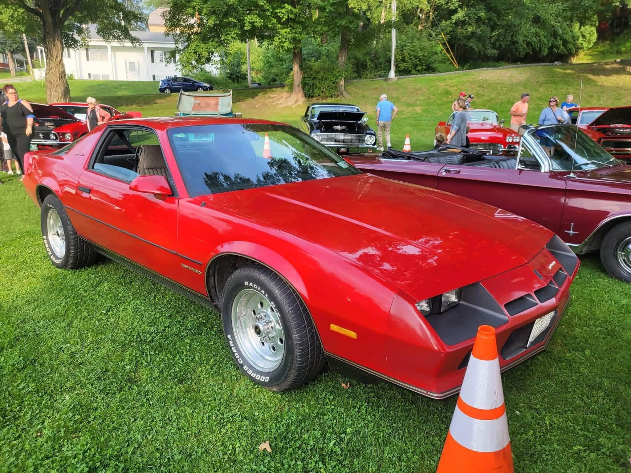 A red Camaro parked on the grass among a row of classic cars at the Wood Park car show.