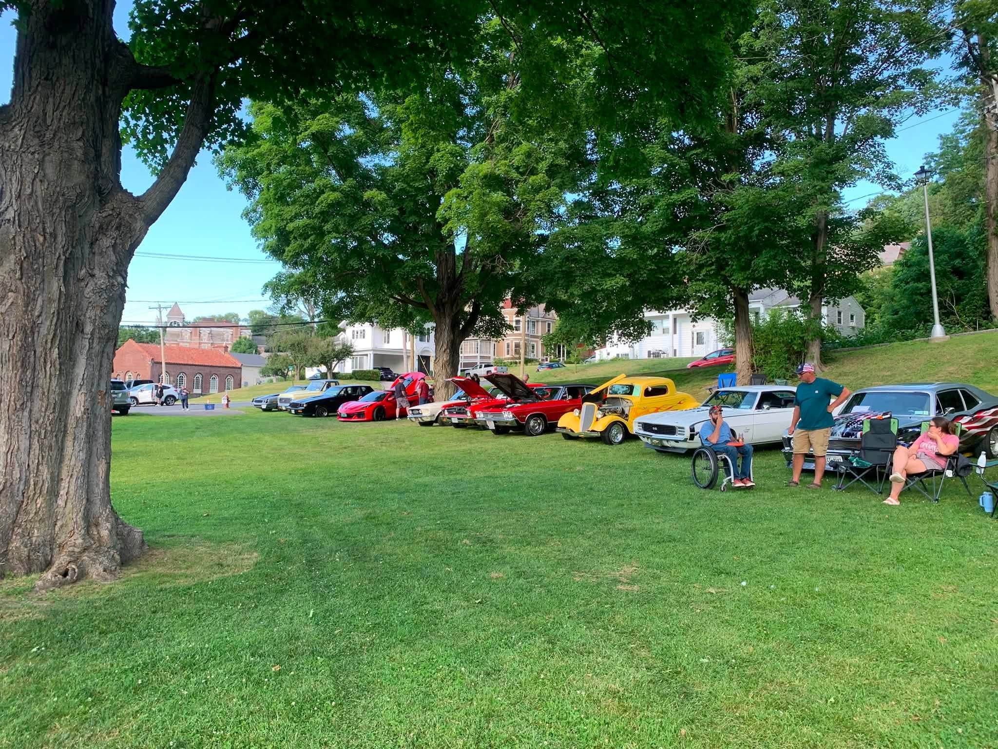 Show cars parked in the shade of the big maples at Wood Park on car show night.