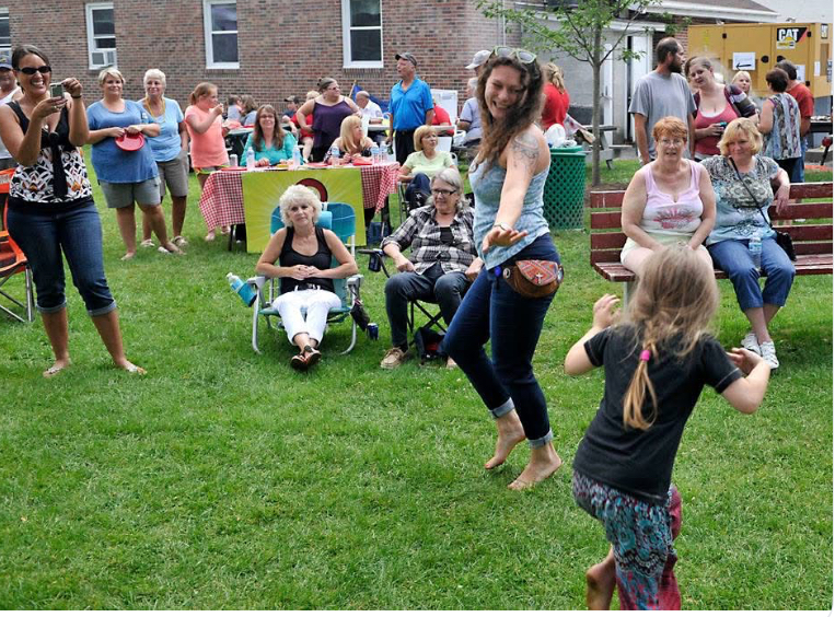 A woman dancing barefoot on the lawn with a little girl while neighbors look on.