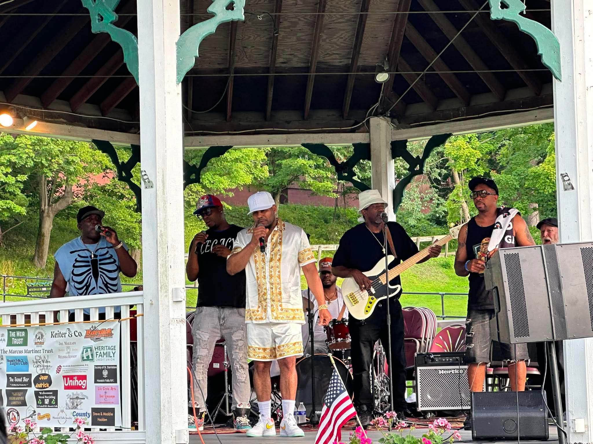 Legacy Music Group lined up across the gazebo stage with the 2026 sponsor banner on the railing.