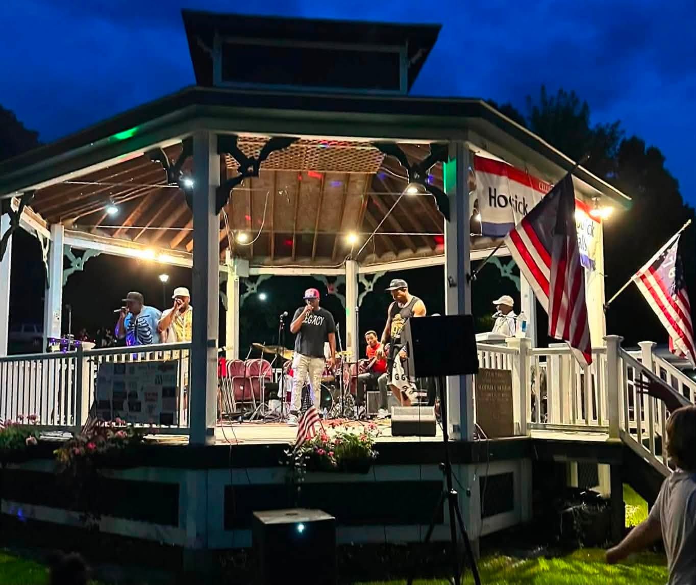 A night set at the gazebo, stage lights on and flags waving as the band plays into the dark.