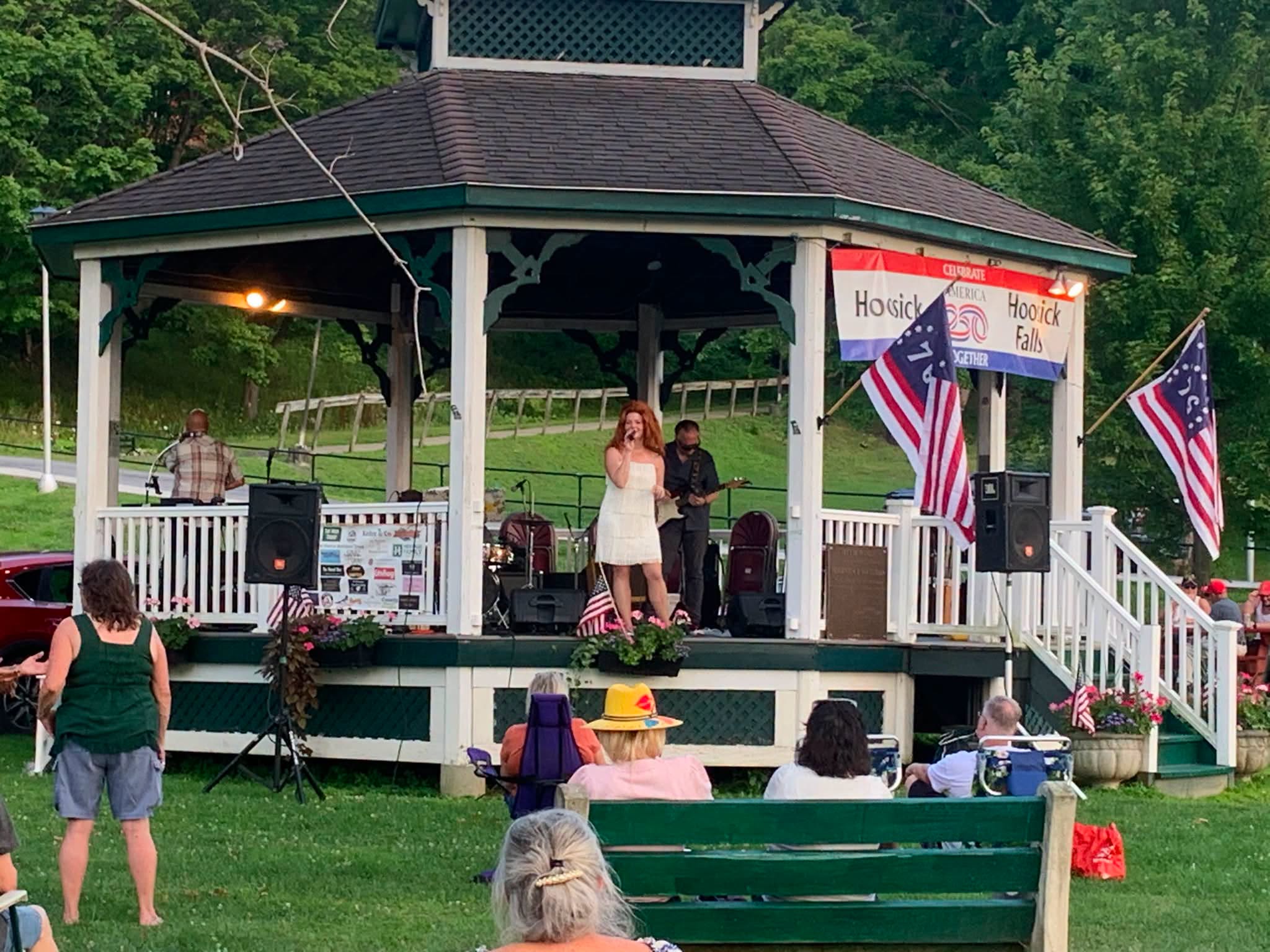 Miss Paula singing at the Wood Park gazebo in a white fringe dress as neighbors watch from the lawn.